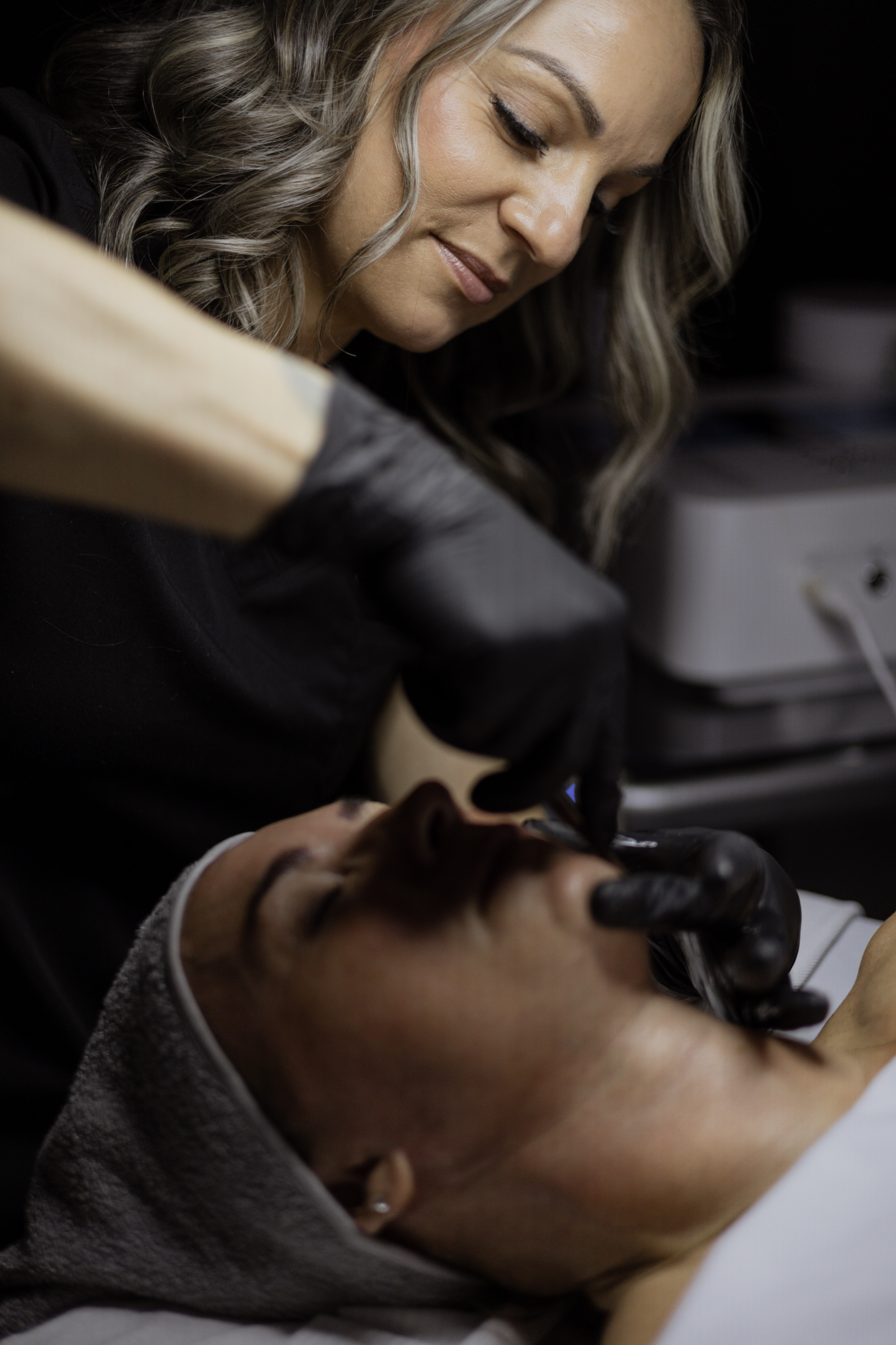 A skincare professional administering a treatment to a client, both are in a spa setting. The client is reclined with a towel on her head, while the professional focuses on the client's face.