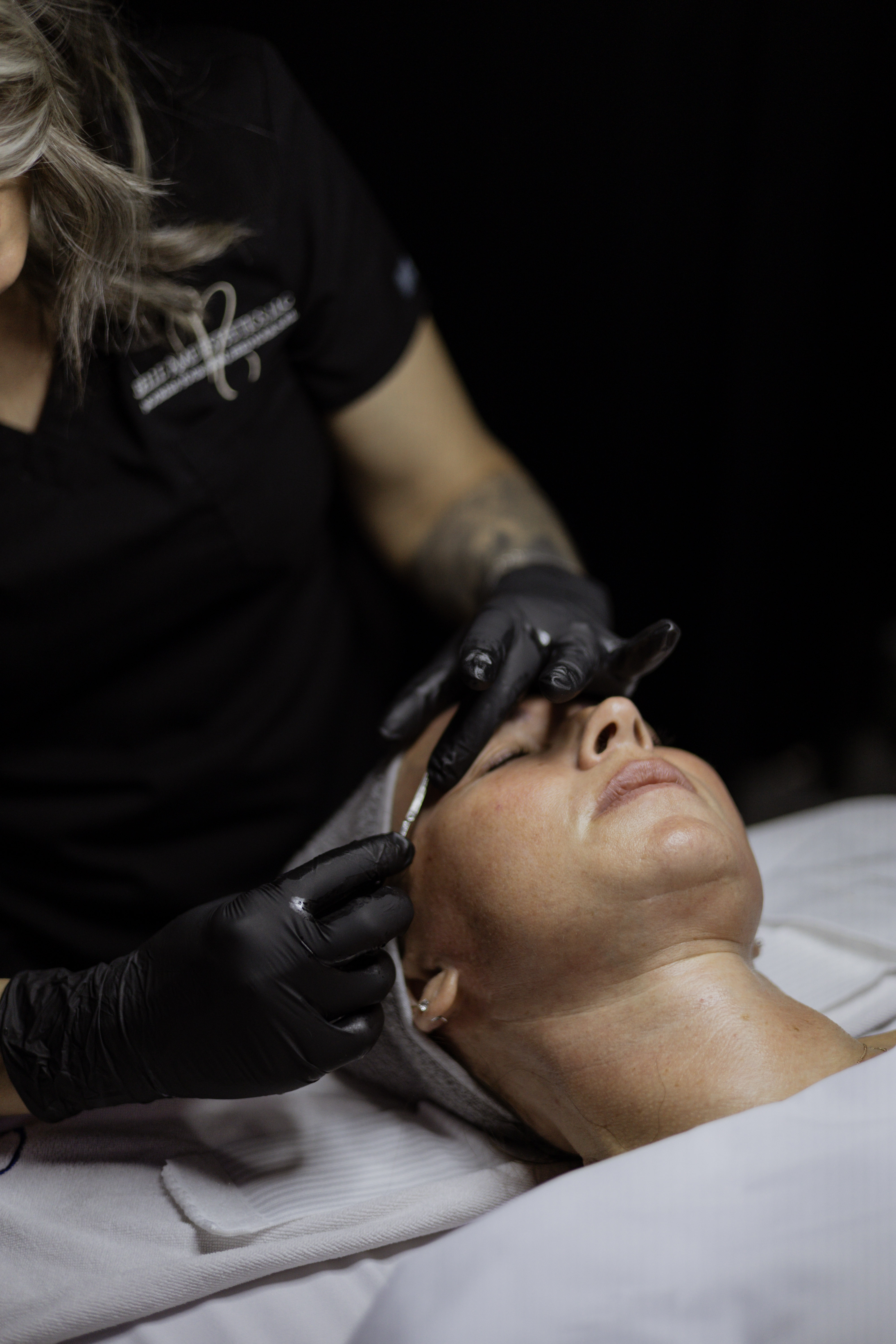 A skincare professional wearing black gloves performs a facial treatment on a client lying on a treatment table, with a relaxed expression.