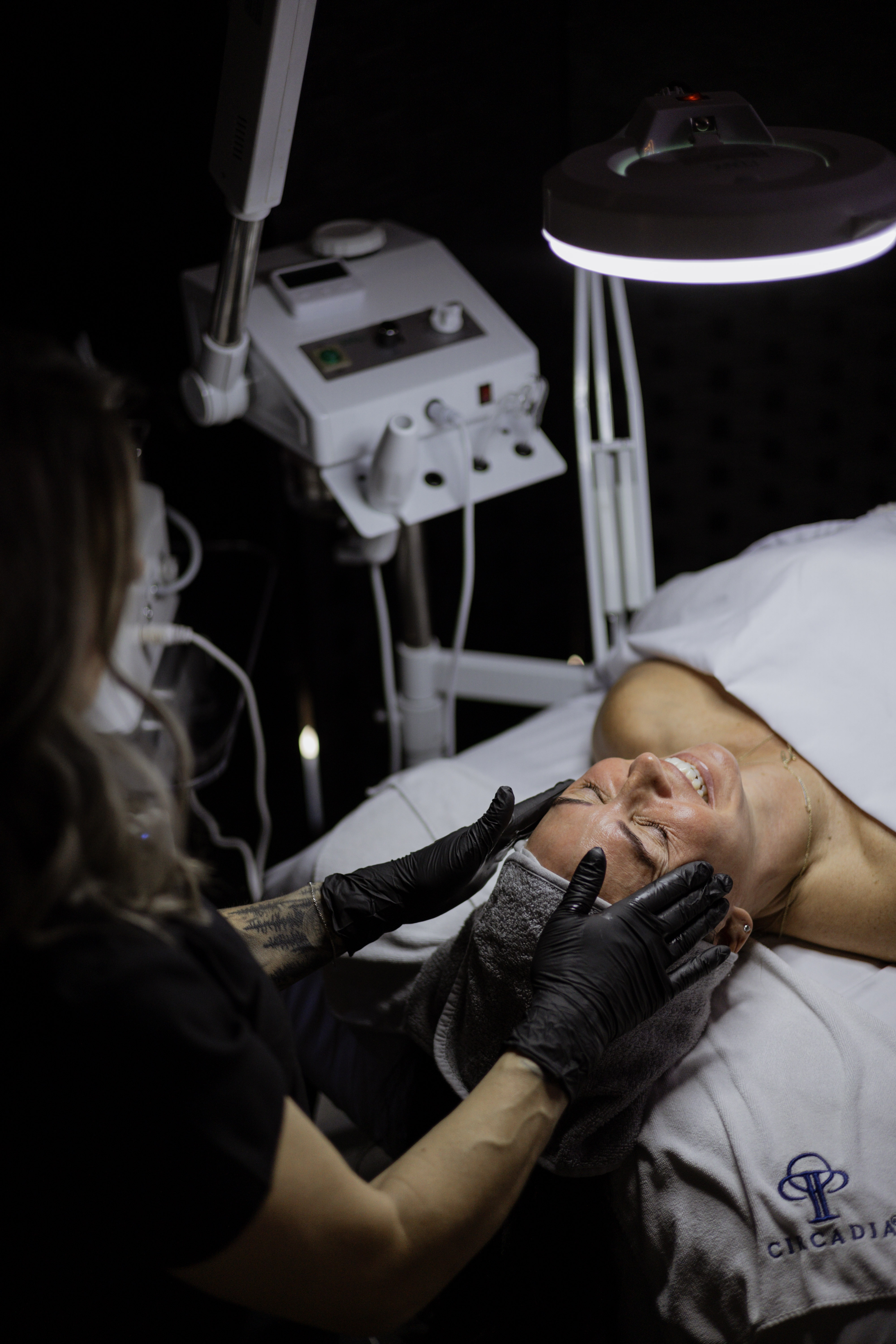 A skincare professional performing a facial treatment on a smiling client in a dimly lit spa environment, surrounded by skincare equipment.