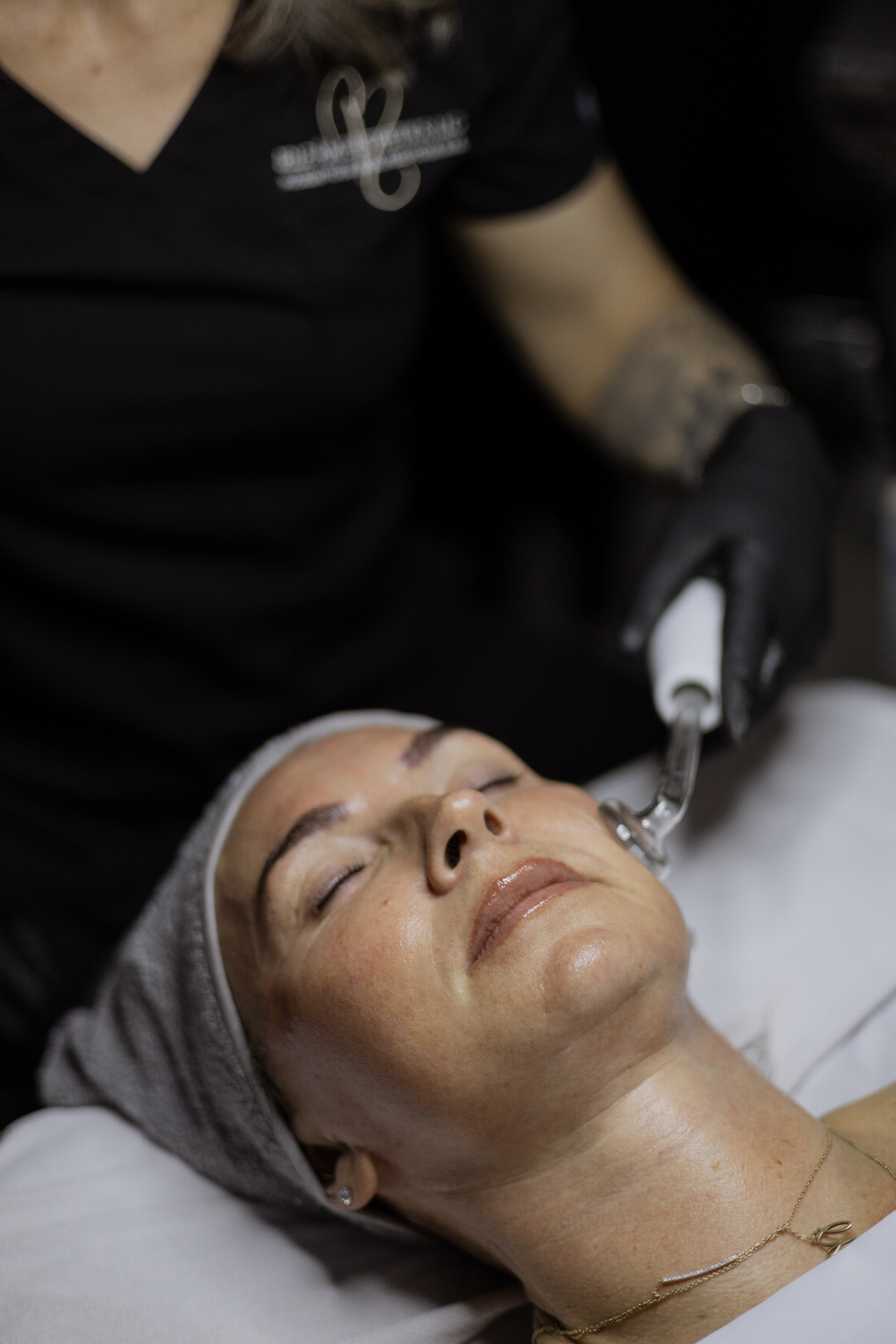 A woman receiving a facial treatment, lying back with her eyes closed while a practitioner uses a skincare device on her face.